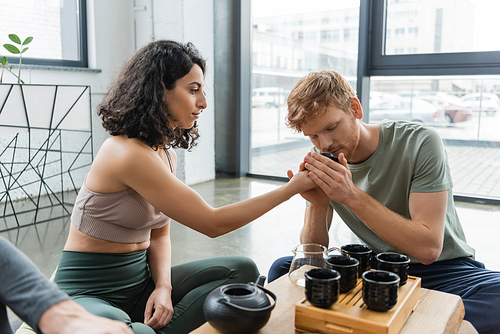 redhead man smelling puer tea near curly middle eastern woman near Chinese teapot and cups