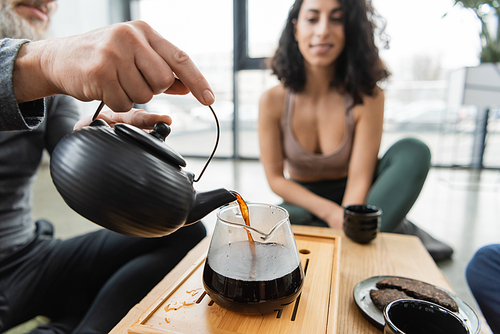 middle aged man pouring brewed puer tea into glass jug near middle eastern woman on blurred background