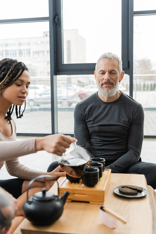 african american woman with dreadlocks pouring puer into traditional tea cups near middle aged man