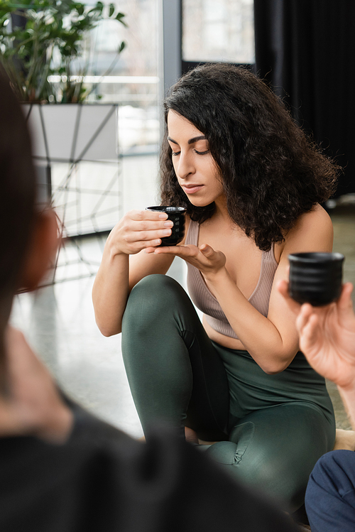 curly middle eastern woman smelling puer tea near people in yoga studio
