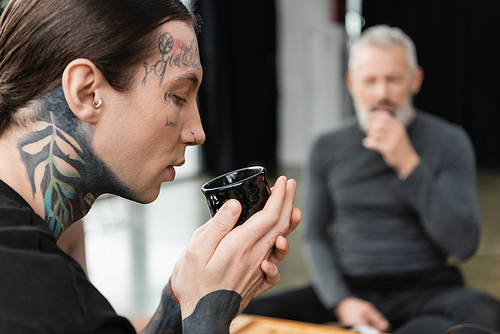 side view of tattooed man holding Chinese cup with brewed puer tea near middle aged man on blurred background