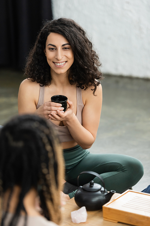 happy middle eastern woman with curly hair holding Chinese cup during tea ceremony in yoga studio
