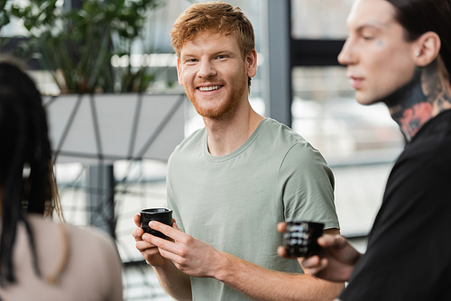 cheerful man with red hair holding japanese cup with puer tea near people in yoga studio