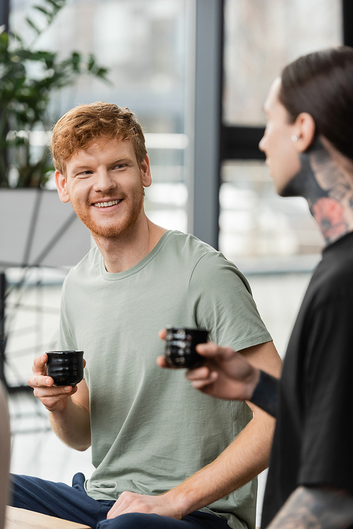 positive man with red hair holding Japanese cup with puer tea near tattooed friend in yoga studio