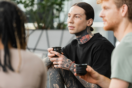 tattooed man holding Japanese cup with tea near people on blurred foreground