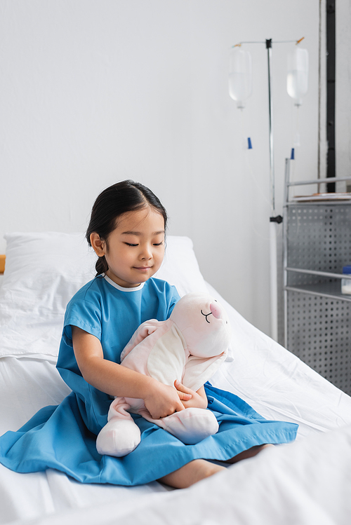 smiling asian girl sitting on bed in hospital ward and hugging toy bunny