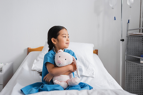 pensive asian girl hugging toy bunny and looking away while sitting on hospital bed