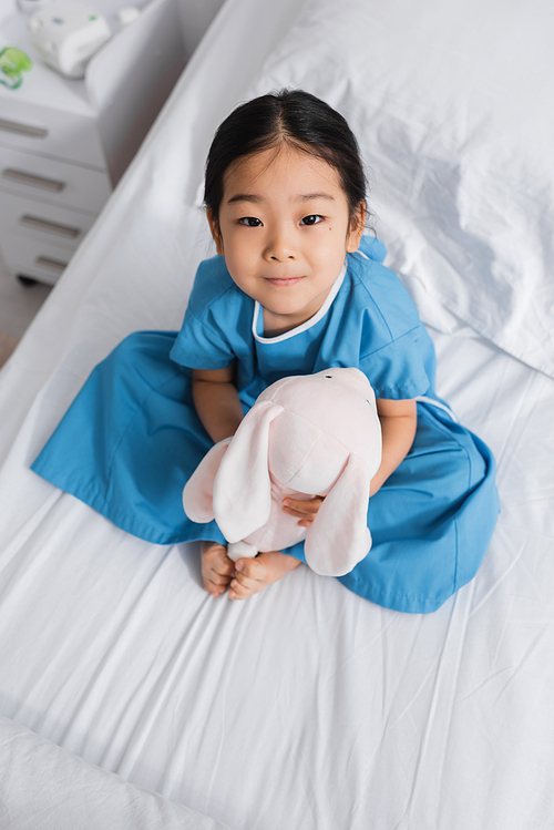 top view of little asian girl sitting on hospital bed with toy bunny and looking at camera