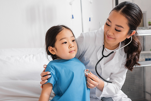 joyful doctor examining little asian girl with stethoscope in pediatric clinic