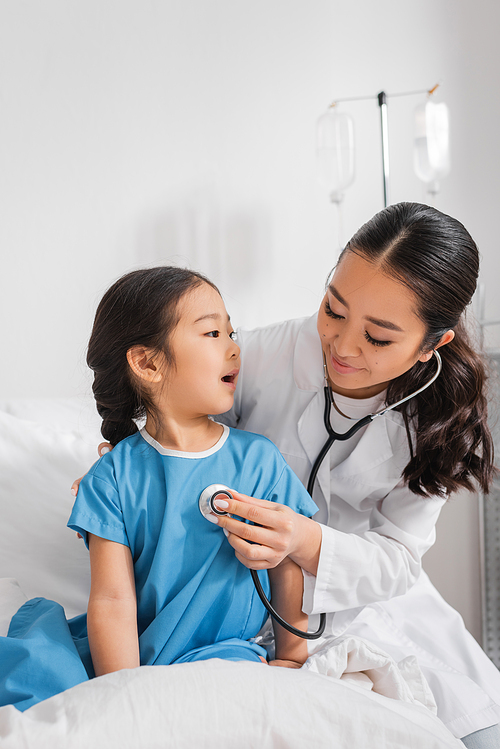 asian girl talking to smiling pediatrician with stethoscope in hospital ward