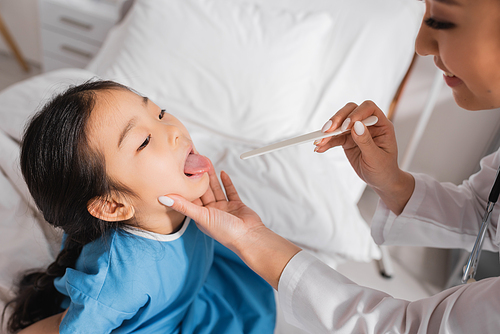 asian girl opening mouth near smiling doctor with tongue depressor in hospital ward