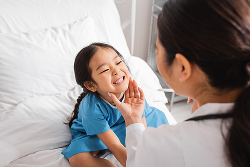 little asian girl showing teeth to blurred doctor with tongue depressor in hospital ward