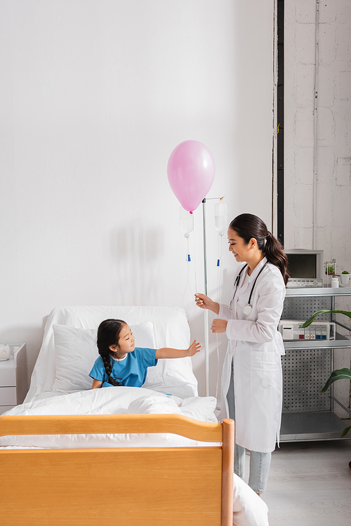 smiling doctor in white coat standing with festive balloon near asian girl on hospital bed