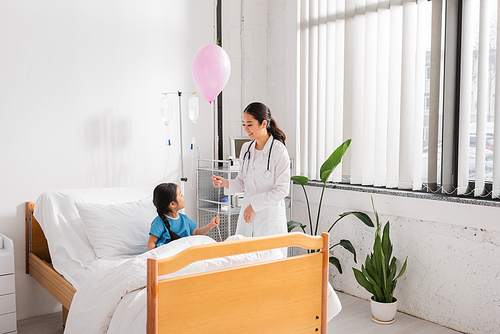 smiling asian doctor holding festive balloon near girl sitting on bed in modern hospital ward