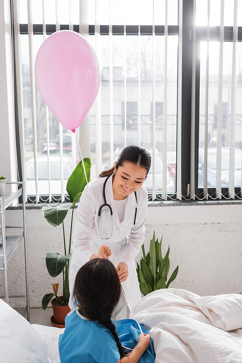 joyful asian doctor giving festive balloon to little patient in pediatric hospital