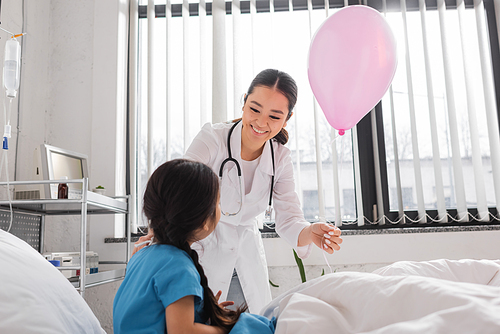 young asian pediatrician with festive balloon smiling near girl sitting on bed in hospital ward