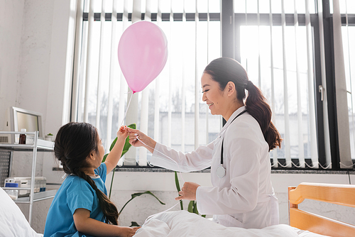 side view of happy asian doctor holding festive balloon near little girl in hospital ward
