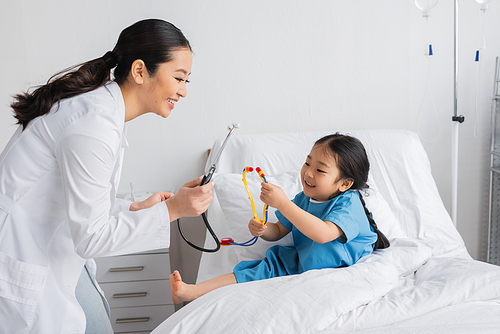 side view of cheerful asian girl showing toy stethoscope to smiling doctor in hospital ward
