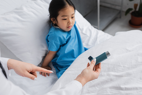 doctor pointing at inhaler near discouraged asian girl sitting on hospital bed