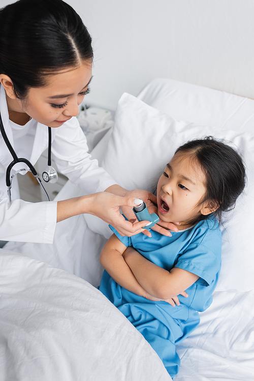 young pediatrician holding inhaler near asian girl with open mouth and folded arms on hospital bed