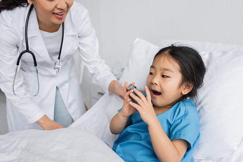 little asian girl with open mouth holding inhaler near doctor in pediatric clinic