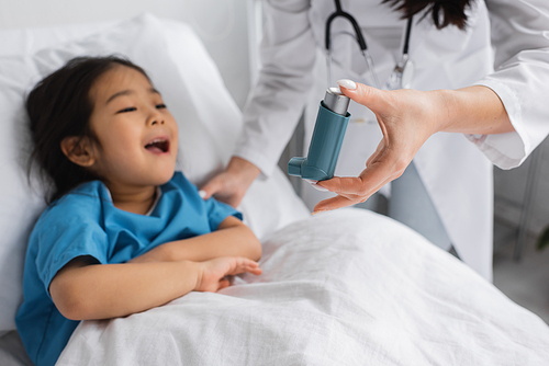 blurred pediatrician holding inhaler near asian child opening mouth on hospital bed