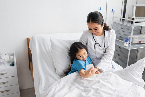 carefree child taking selfie on smartphone with smiling asian doctor in hospital ward