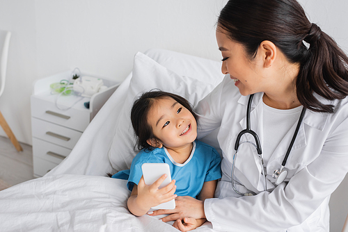 cheerful asian girl looking at young pediatrician and taking selfie on mobile phone in clinic