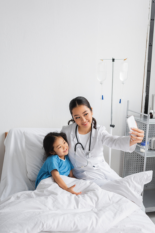 happy doctor in white coat taking selfie with positive asian child in hospital ward
