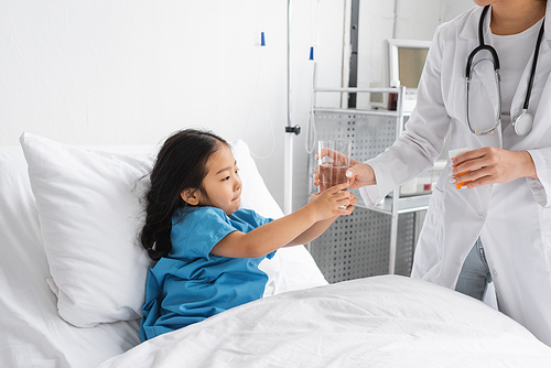 doctor in white coat holding pills container and giving glass of water to asian girl on hospital bed