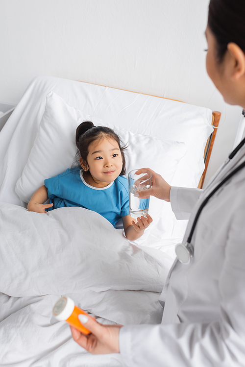 cheerful asian child taking glass of water from doctor holding pills container in hospital ward