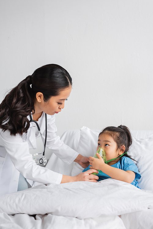 young doctor helping asian girl breathing in oxygen mask on bed in hospital ward