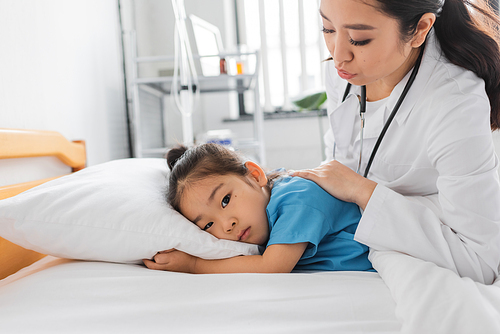 young asian doctor calming sad child lying on bed in clinic