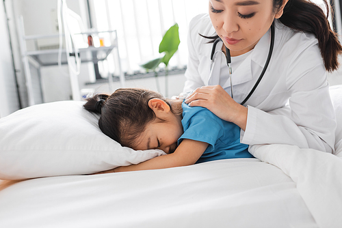 asian doctor calming depressed child obscuring face with pillow on hospital bed