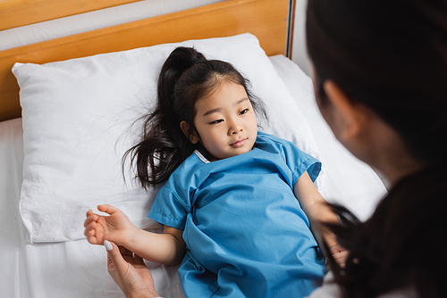 high angle view of little asian girl lying on hospital bed near blurred doctor