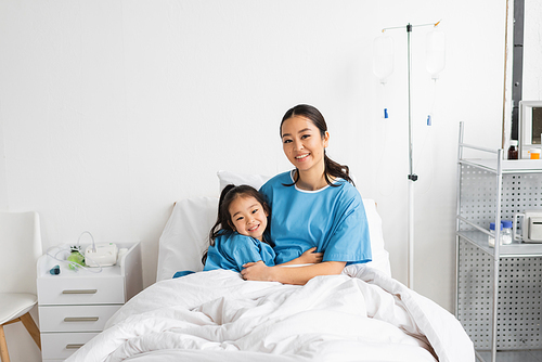 cheerful asian mother and child embracing and looking at camera on bed in hospital ward