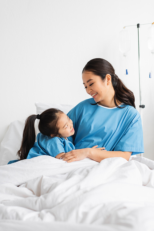 happy asian mother and daughter embracing and looking at each other on hospital bed
