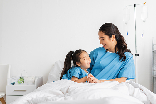 young asian woman holding hands of smiling daughter on bed in hospital ward