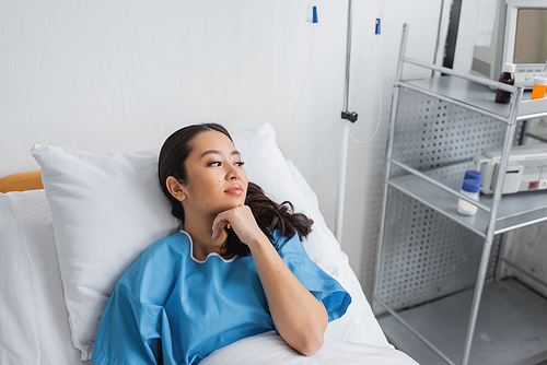 dreamy asian woman holding hand near chin and looking away on bed in hospital ward