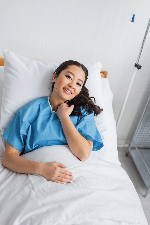 high angle view of pleased asian woman looking at camera while lying on bed in hospital ward