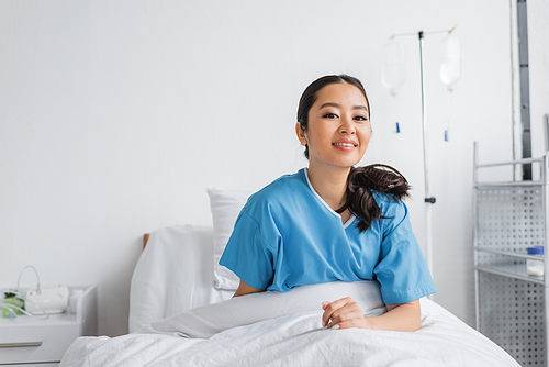 cheerful asian woman sitting on bed in hospital ward and smiling at camera