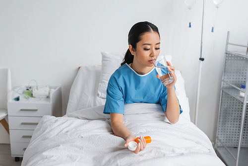 young asian woman drinking water and holding pills container while sitting on bed in hospital ward