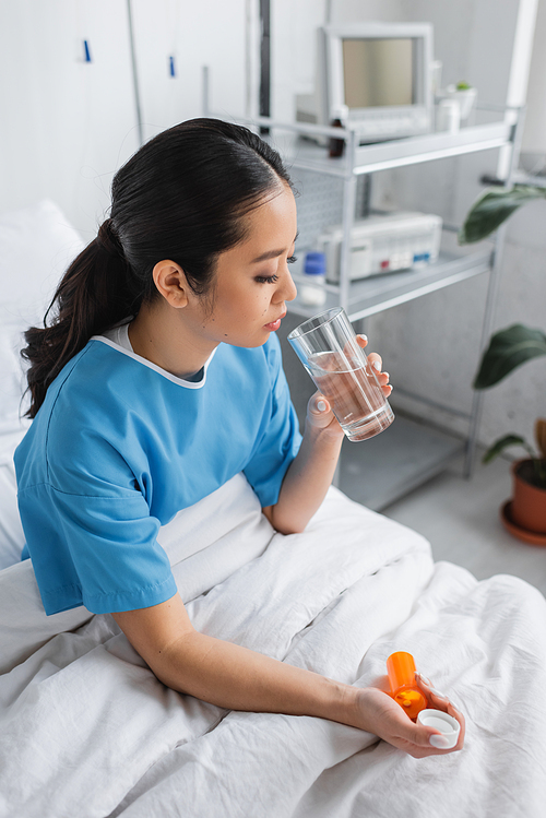 brunette asian woman in hospital gown sitting on bed with glass of water and pills container