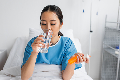 young asian woman drinking water while holding pills container on bed in clinic