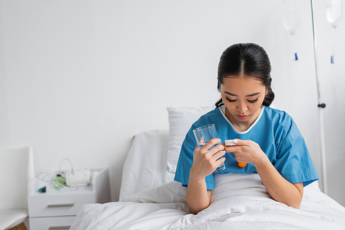 young asian woman looking in pills container while holding glass of water on hospital bed
