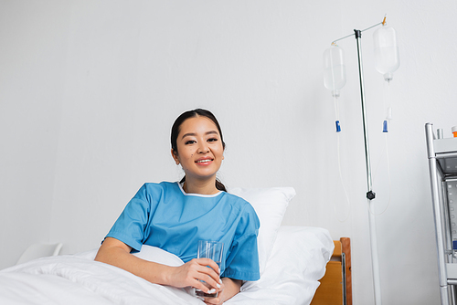 cheerful asian woman holding glass of water and looking at camera on hospital bed