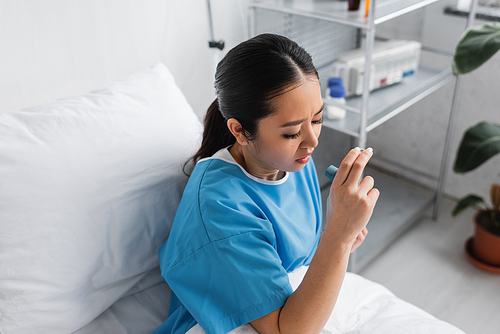 high angle view of sick asian woman holding inhaler while sitting on bed in hospital ward