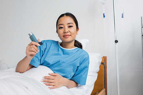 positive asian woman smiling at camera while holding inhaler on bed in hospital ward