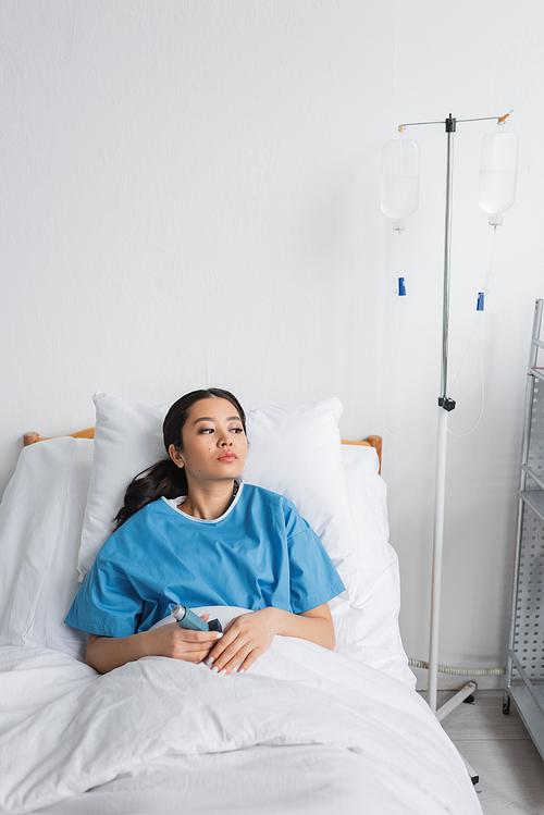 thoughtful asian woman holding inhaler and looking away on bed in hospital ward