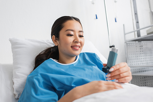 happy asian woman in hospital gown lying on bed in clinic and looking at inhaler